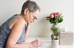Older woman sitting at home, journaling at a table with a tissue box and flower arrangement