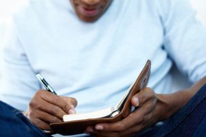 Young man wearing light blue. long-sleeved shirt as he sits and writes in a journal