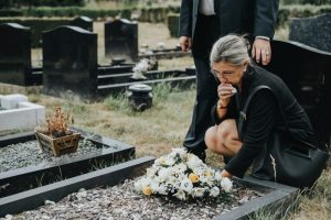 GRay-haired woman kneeling at gravesite, flowers placed on grave with a tissues to her face