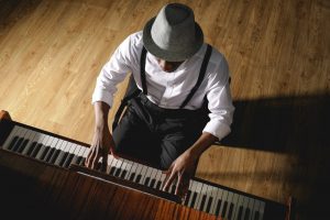 Looking at a man from above as he plays the piano. He wears a hat, white shirt, and dark trousers.