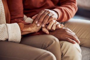 Older man and woman sitting on a couch, holding hands in a comforting way, focus on their hands
