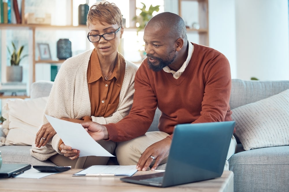 Middle-aged husband and wife sitting on couch at home, reviewing documents together