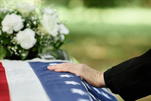 Person standing next to flag-draped casket, hand resting on top of casket