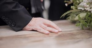 Close-up of hand resting on a casket lid with flower arrangement nearby