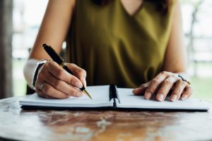 Woman in olive green blouse sitting at a table at home with a pen and checklist in front of her on the table