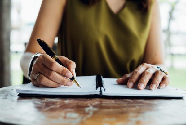 Woman in olive green blouse sitting at a table at home with a pen and notebook in front of her on the table