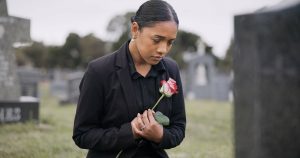 Young woman wearing black kneeling in a cemetery holding a pink memorial rose
