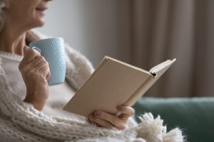 Older woman sitting on couch at home with poetry book, mug, and cozy white blanket