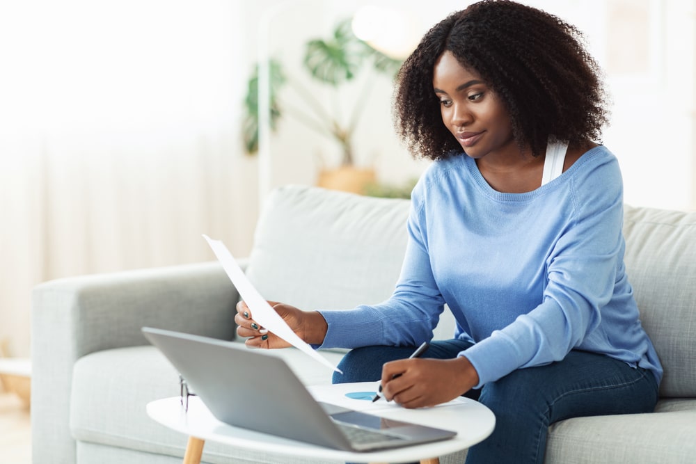 Woman in light blue, long-sleeved shirt siting on couch with computer in front of her, reviewing a piece of paper