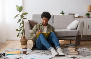 Young man sitting on floor in front of his couch at home, writing poetry in a notebook