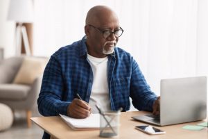 Older man in blue shirt sitting at a table at home with a laptop and notepad in front of him