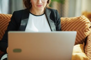 Woman in black blazer sitting on couch and watching livestream on laptop; using technology
