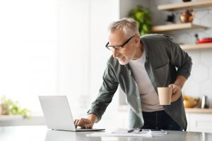 Mature man standing in kitchen with mug in one hand and using the other hand to click on laptop