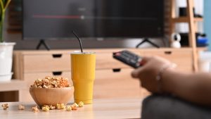 Person sitting on couch at home, holding remote, with popcorn and a drink on a table in front