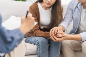 Young husband and wife sitting on a couch holding hands as they speak to a grief therapist