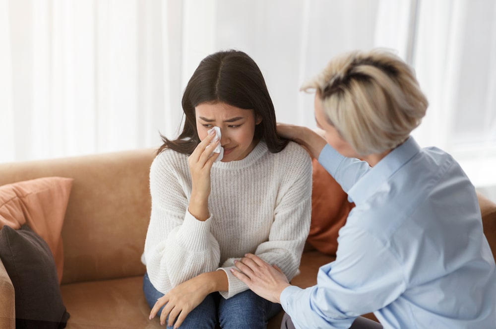 Young woman sitting on couch, wiping eyes with tissues, as grief therapist offers comfort