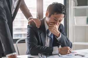 Sad businessman who is sitting at his desk with his hand over his mouth. Co-worker stands nearby with a hand on the sad man's shoulder.