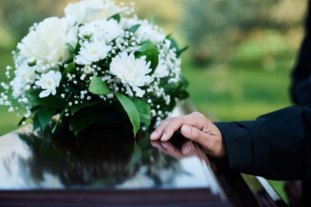 person placing their hand on a coffin with a bouquet of white flowers on top after reading a poem at the funeral