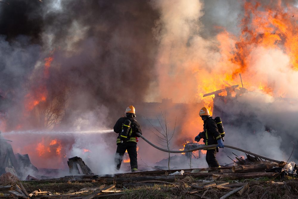 two firefighters holding a water hose, orange flames and smoke in the background