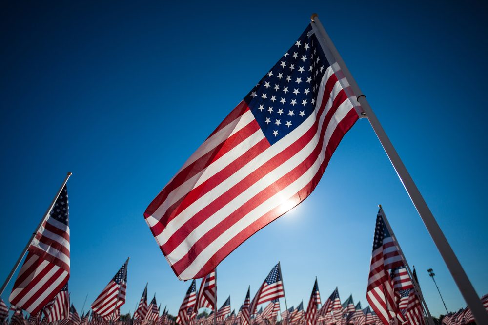 Foreground: American flag backlit by the sun. Background: a grouping of American flags.