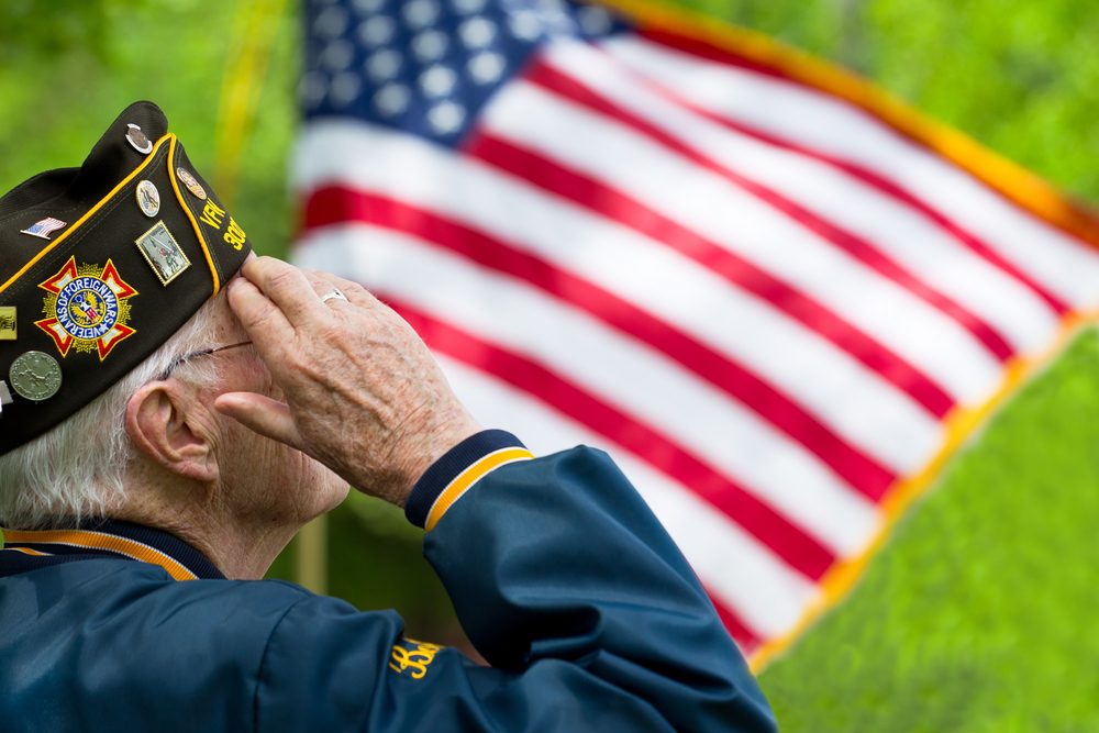 Elderly veteran saluting in foreground, American flag in background
