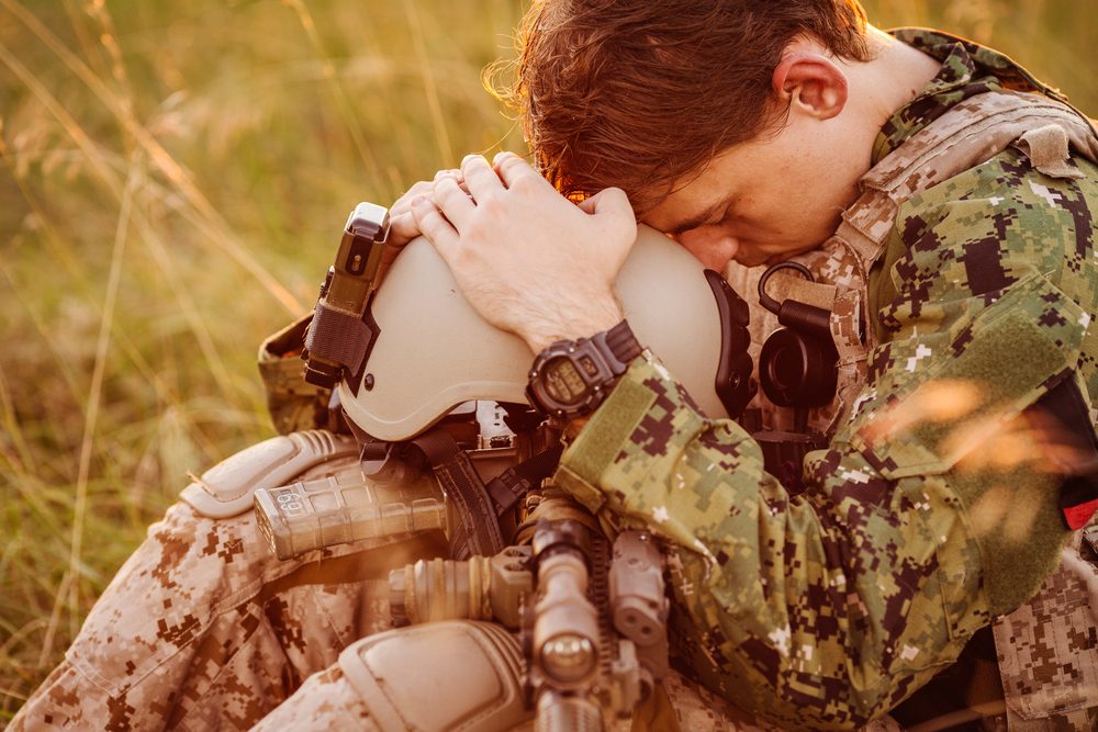 Young male soldier, sitting in a field, helmet in lap, knees drawn up, face pressed to helmet