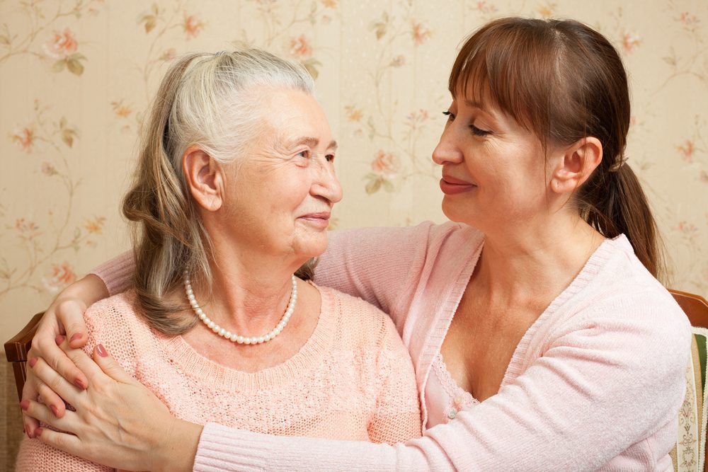 senior mom with her middle-aged daugther, smiling and hugging, wearing light pink blouses.