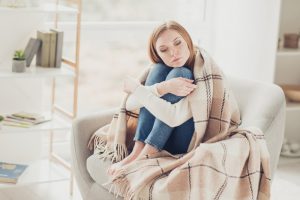 Young woman sitting in a chair alone, wrapped in a blanket, with a sad look on her face