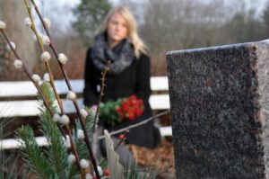 Woman sits near a grave marker with red flowers in her arms, wearing a dark winter coat