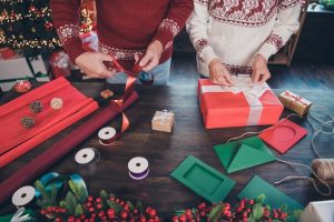 Two people wrapping Christmas gifts together on a table filled with paper, tape, bows