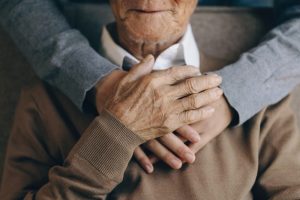 Senior man sitting in a chair with a loved one's arms wrapped around his shoulders