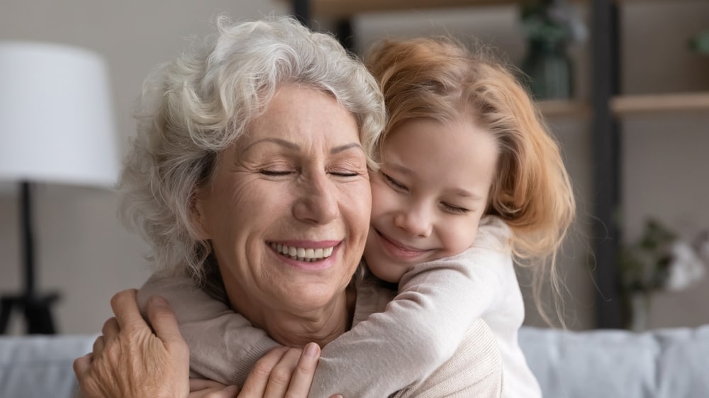 Grandmother and granddaughter sitting on couch and hugging with smiles on their faces