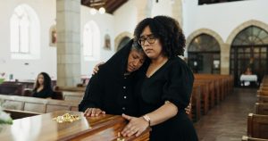 Two women comforting each other at a visitation as they stand next to a casket