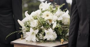 Focus on two pallbearers carrying front of wooden casket with white flower spray resting on lid