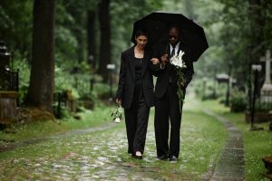 Two people walking through cemetery together with an umbrella and flowers