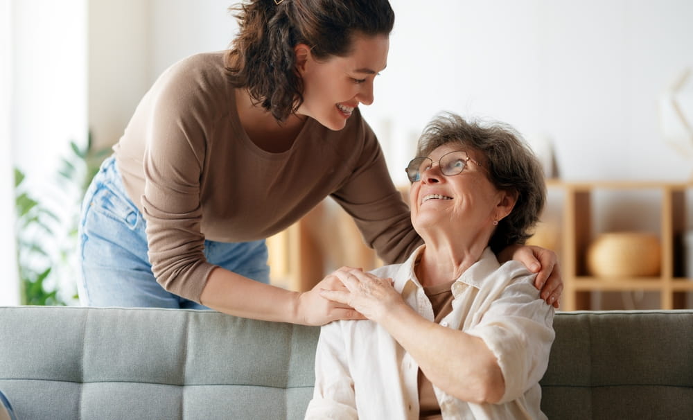 Senior woman sitting on couch and smiling up at adult daughter