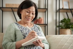 Woman in light blue shirt sitting on couch and writing in gratitude journal