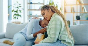 Two female friends sitting on couch, one offering comfort to her crying friend