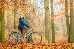 Woman in warm coat riding a bike through a forest filled with fall leaves and barren trees
