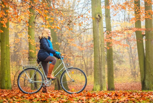 Woman in warm coat riding a bike through a forest filled with fall leaves and barren trees