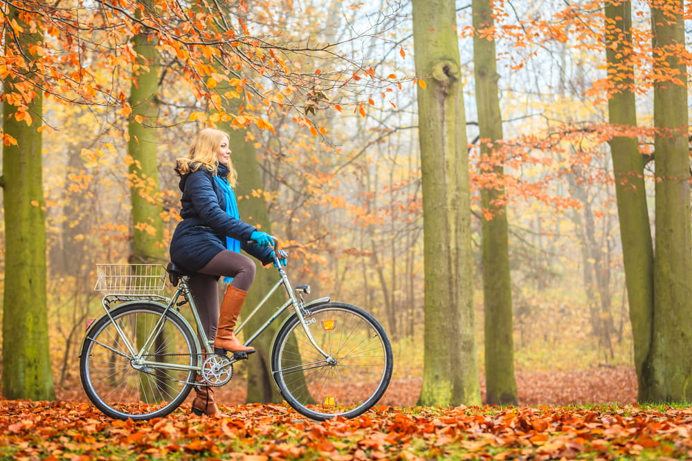 Woman in warm coat riding a bike through a forest filled with fall leaves and barren trees