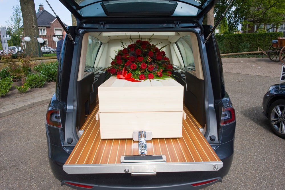 White casket with flower spray of red roses resting in the back of a funeral car