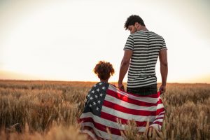 Father and young daughter standing in field with American flag wrapped around them