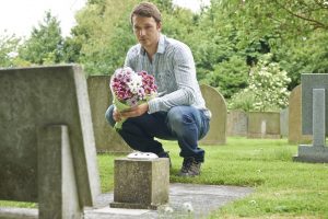 Shows a man leaving flowers at a loved one's memorial marker