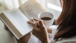 Woman sitting by window with a cup of coffee while reading a book