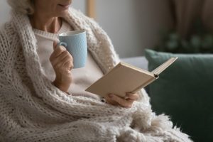 Older woman wrapped in blanket sitting on couch and reading a book