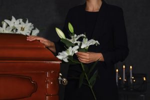 Woman dressed in black standing next to casket with one hand on the casket and the other holding white lilies