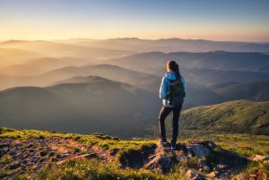 Girl on mountain peak with green grass looking at beautiful mountain valley in fog at sunset in summer.