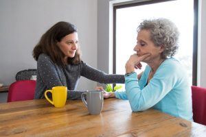 Younger woman comforting older woman by placing hand on shoulder and offering hot tea