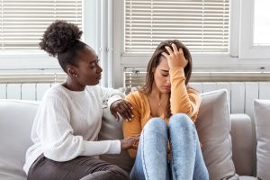 Two young, female friends sitting on a couch, one sad while the other offer support